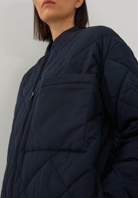 Vrouw in een marineblauwe gewatteerde jas met een grote voorzak en een zwarte ribbelkraag, staand tegen een effen lichte achtergrond.