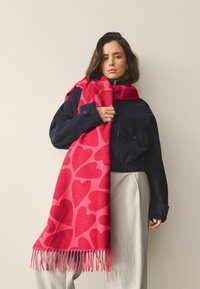 Bufanda roja con patrón de corazones rosas, bordes con flecos, combinada sobre una chaqueta oscura y pantalones de color claro. Textura suave, diseño informal.