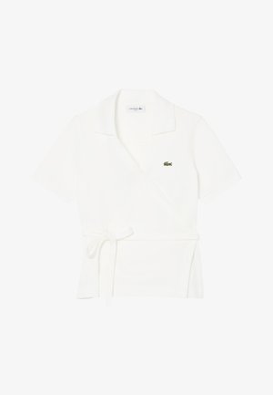 Polo blanco de manga corta tipo wrap con cinturón para anudar en la cintura y pequeño logo de cocodrilo verde en el pecho.