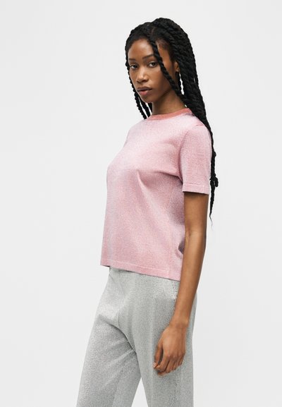 Femme aux longs cheveux torsadés portant un haut rose à manches courtes et un pantalon gris clair, debout devant un fond blanc uni.