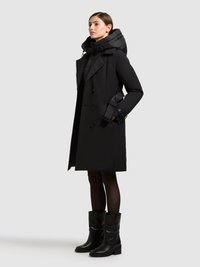 Černý dvouřadý trench coat s vypolstrovanou kapucí, texturovanou tkaninou a nastavitelným páskem na manžetách, nošený přes černou přiléhavou vnitřní vrstvu a černé boty.