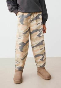 Pantalons à motif camouflage beiges, verts et gris, dotés d'une coupe décontractée et d'une ceinture élastique, associés à des bottines marron.