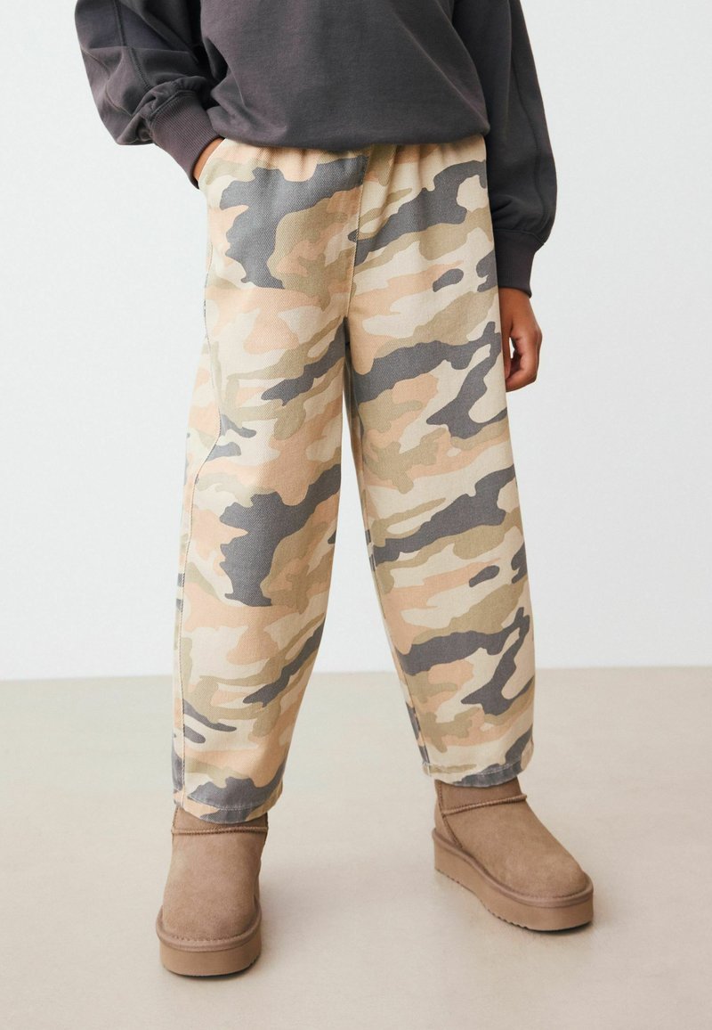 Pantalons à motif camouflage beiges, verts et gris, dotés d'une coupe décontractée et d'une ceinture élastique, associés à des bottines marron.