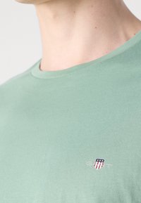 Mintgrøn t-shirt med rund hals, lavet af glat stof, med et lille broderet logo med et skildedesign på venstre bryst.