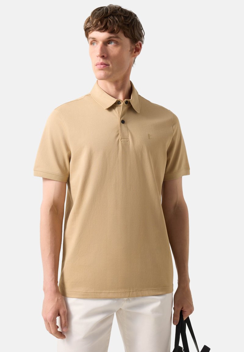 Jonge man met een beige polo met korte mouwen en witte broek, houdt een zwarte tas in zijn rechterhand en kijkt naar links.