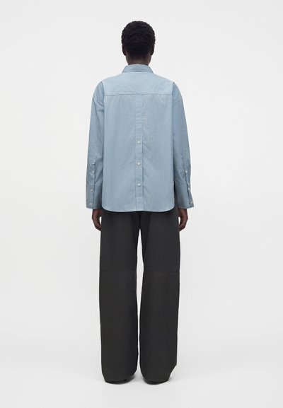 Personne de dos portant une chemise bleu clair à manches longues avec des boutons dans le dos et un pantalon large, sombre et ample.