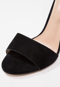 Sandale noire en daim à bout ouvert avec une large lanière avant et une semelle intérieure beige, présentée en gros plan sur un fond blanc.