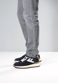 Marineblaue Wildledersneaker mit weißen Akzenten, ausgestattet mit einer dicken cremefarbenen Sohle und schwarzen Schnürsenkeln, getragen zu hellgrauen Jeanshosen.