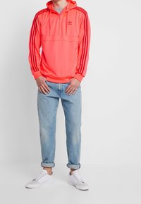 Sweat à capuche orange néon avec un demi-zip, poche frontale et accents rouges à trois bandes. Associé à un jean bleu clair avec revers et des baskets blanches.