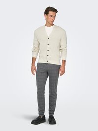 Cardigan en maille beige clair avec un col en V, cinq boutons foncés, associé à un pantalon slim à carreaux gris et des chaussures noires.