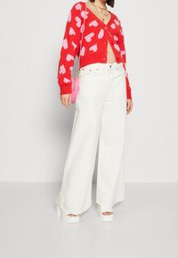 Roter Cardigan mit rosa Herzmustern, Knopfverschluss und langen Ärmeln. Weiße weit geschnittene Hosen und klobige weiße Plateausandalen. Rosa Handtasche.