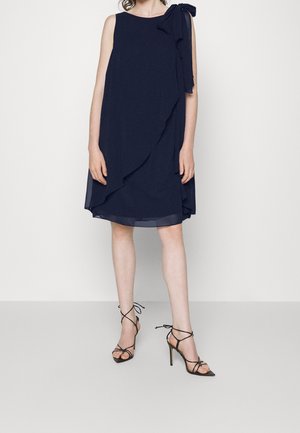 Ärmelloses, marineblaues knielanges Kleid mit asymmetrischem Rüschendetail und Schleife an der Schulter, kombiniert mit schwarzen, hochhackigen Riemchensandalen.