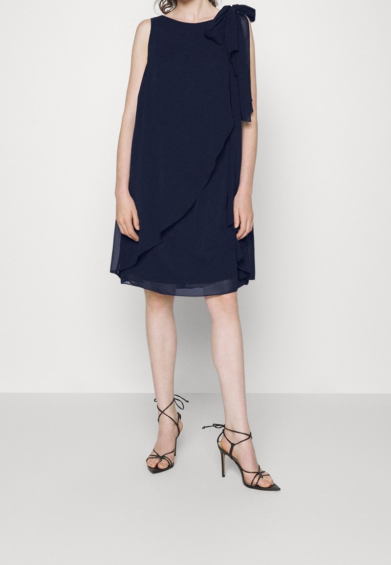 Robe bleu marine sans manches longueur genou avec volant asymétrique et nœud sur l'épaule, portée avec des sandales à talons hauts noires à brides.
