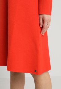 Rotes Wollkleid mit glatter Textur, geradem Schnitt, langen Ärmeln und einem kleinen Logodetail nahe dem Saum.