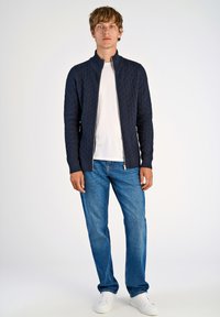 Marinblå zip-up tröja med diamantmönster, ribbad krage och muddar, kombinerad med blå jeans och vita sneakers. Neutral bakgrund.