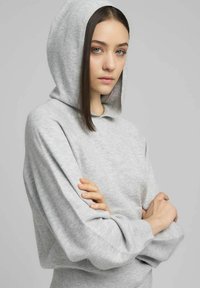 Sudadera con capucha de color gris claro, confeccionada en tela suave, con capucha ajustable mediante cordón, corte holgado y mangas anchas, rematada con un dobladillo acanalado.