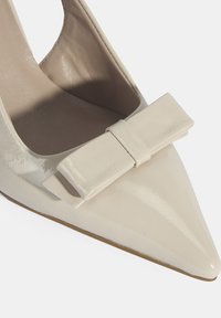 Pointe de talon aigu en cuir verni beige avec un accent de nœud plat sur le devant. Texture lisse et design minimaliste.