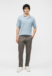 Νέος άνδρας που στέκεται με το ένα χέρι στην τσέπη, φοράει ανοιχτό μπλε κοντομάνικο πουκάμισο, γκρι παντελόνι και λευκά αθλητικά σε μονόχρωμο φόντο.