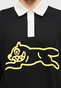 Μαύρο πόλο πουκάμισο με άσπρο γιακά και μεγάλο κίτρινο γραφικό ενός στυλιζαρισμένου γάτου σε θέση άλματος στο μπροστινό μέρος.