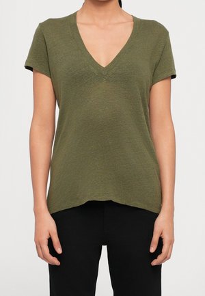 Femme portant un t-shirt vert olive à manches courtes et col en V ainsi qu'un pantalon noir, debout devant un fond clair uni.