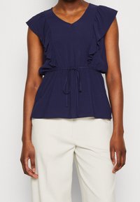 Námořnická modrá blůza s výstřihem do V, volánkovými krátkými rukávy a stahovacími šňůrkami v pase. Vyrobená z měkké tkaniny, s volným střihem a jemnou texturou.