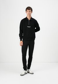 Crna zip-up dukserica s bijelim logotipom, uparena s crnim uskim trapericama i bijelim tenisicama s sivim detaljima. Tkanina izgleda glatko.