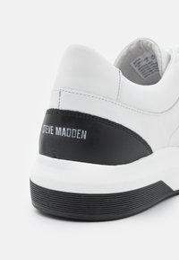 Baskets en cuir blanc avec un panneau de talon en cuir noir. Présente une surface texturée, un branding prominent et une semelle en caoutchouc avec des rainures.