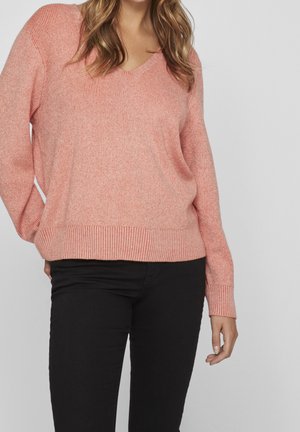 Femme portant un pull en maille rose ample à col en V et un pantalon noir, debout avec une main dans la poche sur un fond gris uni.