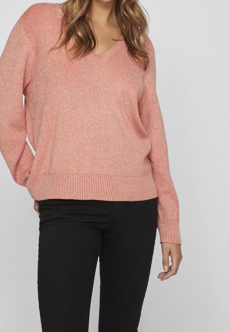 Femme portant un pull en maille rose ample à col en V et un pantalon noir, debout avec une main dans la poche sur un fond gris uni.
