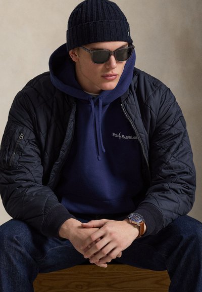 Mladý muž nosiaci tmavomodrú čiapku, slnečné okuliare, tmavomodrú mikinu Polo Ralph Lauren, čiernu prešívanú bundu, džínsy a náramkové hodinky s hnedým koženým remienkom.