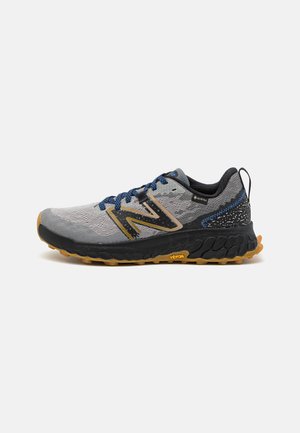Zapatilla de correr gris y negra con parte superior texturizada, cordones azules y suela Vibram. Cuenta con un forro impermeable Gore-Tex y un diseño de logo audaz.