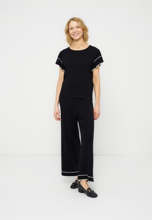 Γυναίκα στέκεται και χαμογελά φοράει μαύρο παντελόνι με φαρδιά μπατζάκια και αντίστοιχο κοντό μανίκι με άσπρη επένδυση, και μαύρα loafers με τακούνι.