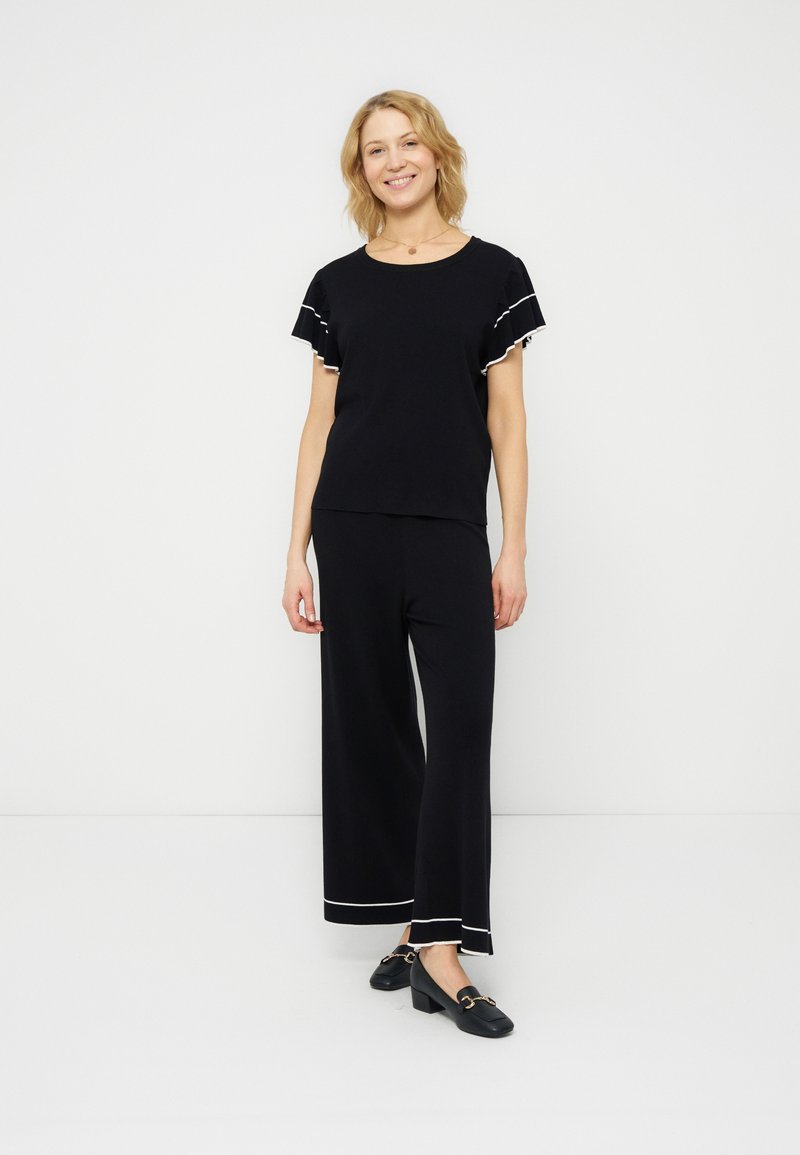 Γυναίκα στέκεται και χαμογελά φοράει μαύρο παντελόνι με φαρδιά μπατζάκια και αντίστοιχο κοντό μανίκι με άσπρη επένδυση, και μαύρα loafers με τακούνι.