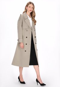 Beige trenchcoat met dubbele rij knopen, brede kraag en schouderepauletten, gecombineerd met een zwarte rok tot op de knie en zwarte hakken.
