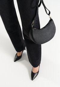 Borsa a spalla in pelle nera con finitura texturizzata, forma curva e chiusura con zip. Indossata con scarpe col tacco nere e jeans scuri.