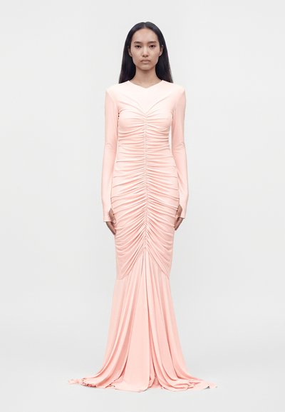Femme aux longs cheveux foncés portant une robe rose pâle à manches longues, froncée, avec une traîne au sol, debout devant un fond uni.