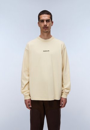 Hombre con camisa beige de manga larga con texto "NAPAPIJRI" y pantalones marrón oscuro, de pie frente a un fondo liso color gris claro.