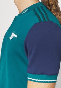 Maillot bleu sarcelle et bleu marine avec un col à boutons, comportant trois rayures bleu marine sur l'épaule et un logo sur la poitrine. Tissu texturé.