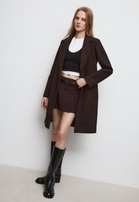 Bruine wollen jas over een zwarte crop top en bordeauxrode geplooide rok, gecombineerd met glanzende zwarte kniehoge laarzen. Eenvoudig ontwerp met een klassieke silhout.