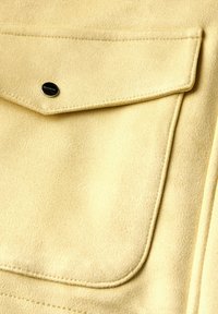 Tissu jaune texturé avec une poche à rabat, doté d'un bouton-pression noir. Matériau doux, semblable à du daim, avec des détails de couture soignés.