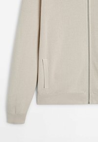 Cardigan beige en maille avec fermeture éclair, manches longues, poignets et ourlet côtelés, et poche fendue verticale sur le côté.
