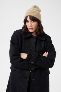 Cappotto nero texturizzato con grandi bottoni, dotato di colletto e tasche frontali. Indossato con un berretto lavorato a maglia beige con una striscia bianca.