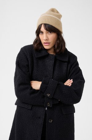Cappotto nero texturizzato con grandi bottoni, dotato di colletto e tasche frontali. Indossato con un berretto lavorato a maglia beige con una striscia bianca.