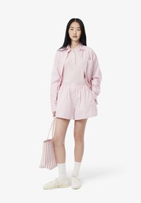 Bleda roza polo majica, lahka roza in bela črtasta oversize srajca, usklajeni šortki in bele športne čevlje, z črtasto torbo.