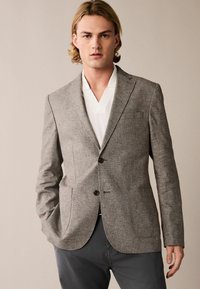 Grau strukturierter Blazer mit zwei Fronttaschen, Reverskragen und zwei Knöpfen. Getragen über einem weißen Hemd mit Kragen, dazu graue Hosen.