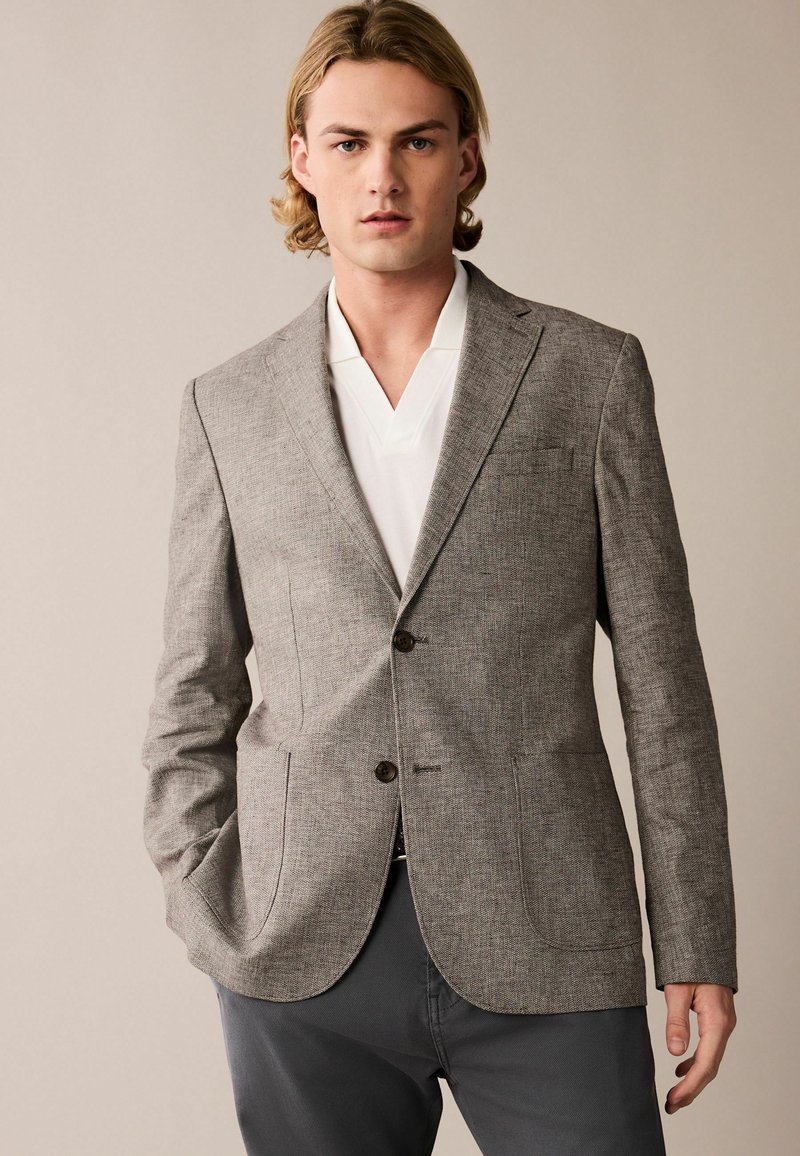 Grau strukturierter Blazer mit zwei Fronttaschen, Reverskragen und zwei Knöpfen. Getragen über einem weißen Hemd mit Kragen, dazu graue Hosen.