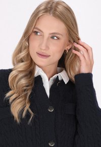 Cardigan navy a maglia cable-knit con bottoni argento, con tasca frontale e indossato sopra una camicia bianca con colletto. Texture liscia e design strutturato.