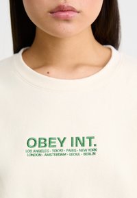 Cream-colored sweatshirt with a round neckline featuring green embroidered text that reads "OBEY INT." and city names underneath in smaller text.