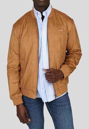 Homme portant une veste aviateur en daim marron clair sur une chemise à rayures bleu clair et blanches avec un jean bleu, posé devant un fond gris.