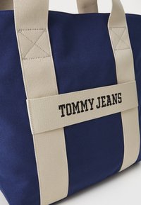 Marinblå canvas tote-väska med beige remmar som har "TOMMY JEANS" broderat i svart. Fyrkantig form, robust konstruktion, synliga sömdetaljer.